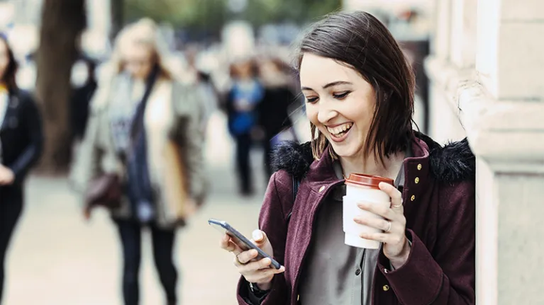 Woman outside, smiling, looking at phone and with a coffee cup in her hand