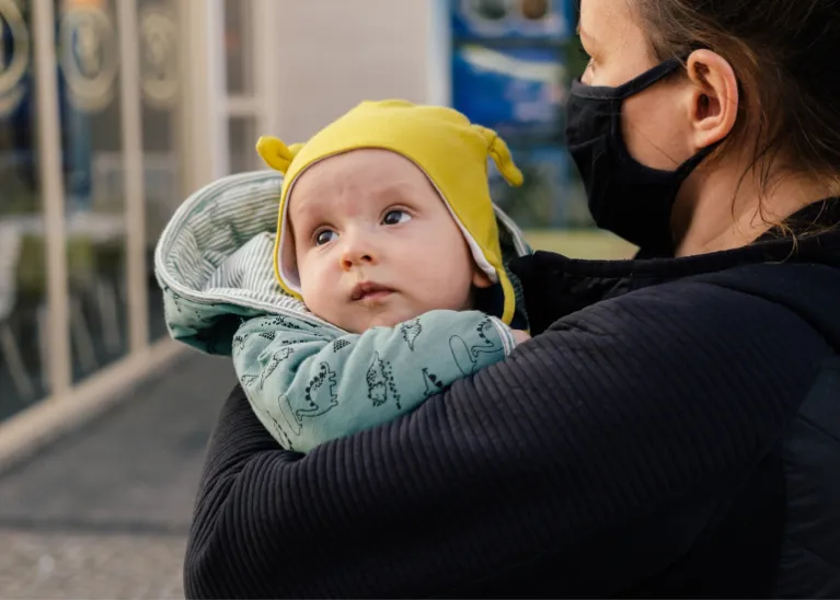 Small baby wearing a yellow hat with a mother wearing a face mask